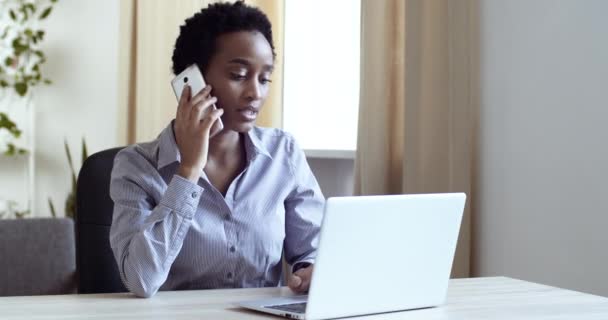 Σοβαρή εστιασμένη Αφροαμερικανίδα επιχειρηματίας που μιλάει στο κινητό τηλέφωνο καλώντας στο γραφείο στη δουλειά συζητώντας με πελάτες ή συναδέλφους που κοιτάζουν το laptop. Επικοινωνία και έννοια της εργασίας — Αρχείο Βίντεο