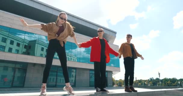 Tři stylové teenageři stojí společně venku proti nedaleké skleněné modré budově, nosí trendy světlé košile a sluneční brýle, drží se za ruce, mávají rukama a rameny, předvádějí taneční prvky — Stock video