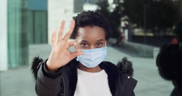 Portret van Afro-Amerikaans meisje positieve vrouwelijke patiënt in medisch beschermend masker staat buiten in de buurt van de stad gebouw toont teken van toestemming symbool van overeenstemming maakt oke gebaar met beide handen — Stockvideo