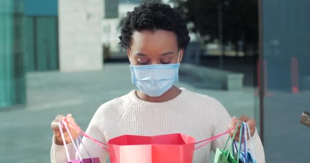 Portrait afroamerikanischer Frau gemischte Rasse Shopaholic Mädchen Shopper Verbraucher trägt medizinische Maske auf dem Gesicht blickt in Farbbeutel mit Einkaufsgeschenken neue Kleidung fühlt Freude Freude Schock Blick in die Kamera — Stockvideo