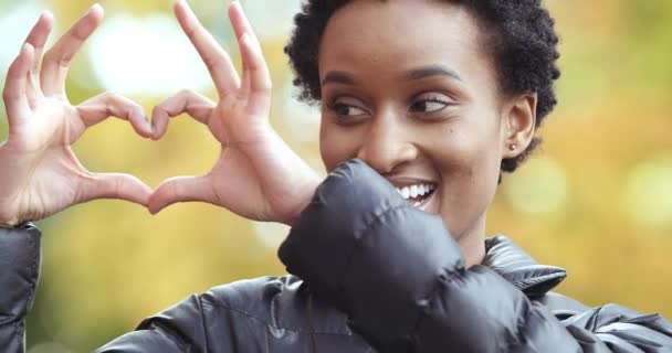 Portré mosolygós érzelmi barátságos etnikai afro-amerikai nő lány áll a szabadban ősszel mutatja a kezét szimbóluma a szeretet jele a béke romantika, hogy ujjak szív alakú mosolygós néz kamera — Stock videók