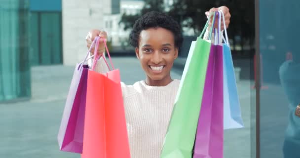 Χαμογελώντας μαύρη γυναίκα αφροαμερικανός καταναλωτής κορίτσι στα λευκά στέκεται σε εξωτερικούς χώρους κοντά εμπορικό κέντρο δείχνει σε φωτογραφικές μηχανές αγορές που αγοράζονται σε εκπτώσεις κατέχει τσάντες ψώνια χρωματιστά πακέτα χαρτόνι στα χέρια — Αρχείο Βίντεο