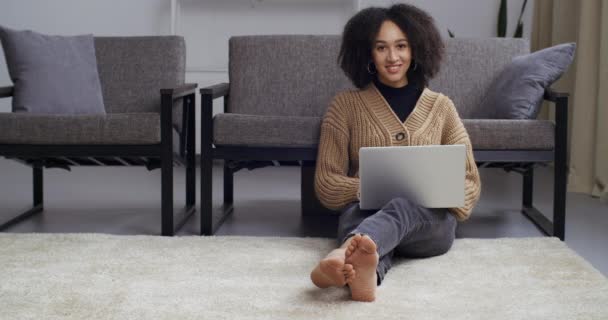 Estudante afro-americano adolescente mista mulher negra sentada no chão em casa perto do sofá trabalhando remoto com laptop olhando para tela datilografando meil estudando online olhando para câmera sorrisos amigável — Vídeo de Stock