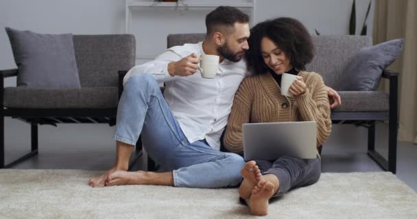 Пара молодоженов муж и жена белый мужчина и афро-американка, сидящие дома на полу возле дивана, пьющие горячий чай кофе из белых чашек, глядя в экран ноутбука смотреть кино серфинг сети — стоковое видео