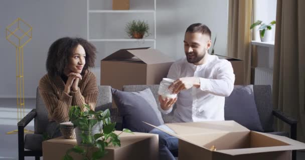 Multietnický pár africká americká dívka a běloch muž sedí spolu na pohovce v pokoji v novém domě, stěhování do bytu, třídění krabic věcí, příjem balíků, chlap dává své ženě dárek — Stock video