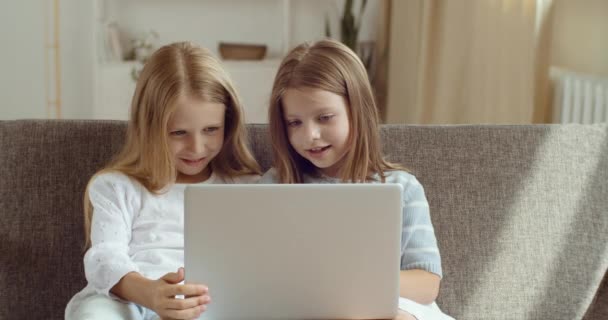 Vista directa de dos hermanas sonrientes mirando el portátil. Retrato de niñas niños usan computadora para ver dibujos animados películas videos divertidos en Internet riéndose juntos sentados en el sofá en casa — Vídeo de stock