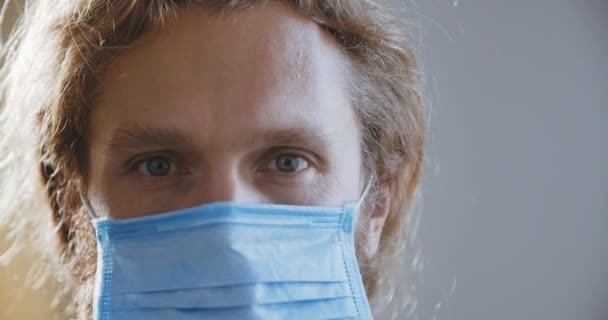 Retrato pandémico de un joven guapo con ojos azules usando máscara médica protectora, concepto de salud y seguridad, cuarentena por coronavirus, protección contra virus. El paciente se para en el hospital mira a la cámara — Vídeos de Stock