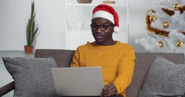 Afro American man black man wears Santas hat sits with laptop on sofa at home near cristmas tree speaks in video chat communicates online in conference with friend family congratulates happy new year — Stock Video