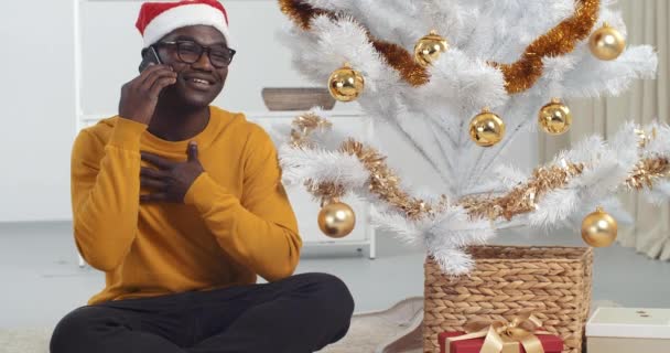 Afrykański Amerykanin siedzi w pokoju z świąteczne dekoracje posiada inteligentny telefon odebrać telefon w Wigilię gratuluje krewnym 2021 cieszyć się zdalną rozmowę z rodziną czuje radosny świętuje Nowy Rok — Wideo stockowe
