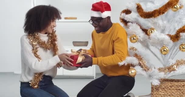 Mixed race couple exchanging present at holiday party in tnw year, husband gives his wife gift box, afro woman gives man new thing, young family friends sitting around Christmas tree at home on floor — Stock Video