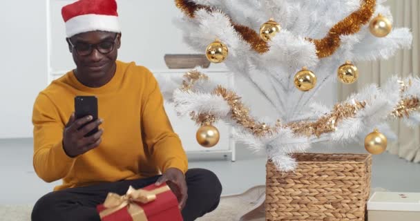 Afro american black man with glasses sits on floor of house near Christmas tree wears Santa Claus hat holds smartphone speaks to camera of mobile phone makes video for blog communicates in online chat — Stock Video