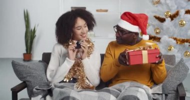 Etnik erkek ve kadın kanepede oturup hediyelerini paylaşıyorlar. Afro-Amerikan çift aile dostları karı koca Noel 'de hediyelerin tadını çıkarıyorlar yeni yılbaşı konseptiyle birlikte.