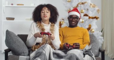Kış tatilinde oyun ve eğlence. Afro-Amerikalı koca ve kıvırcık kadın konsol oynuyorlar. Oyun kağıtlarını tutuyorlar. Kanepede oturup Noel şapkası takıyorlar.