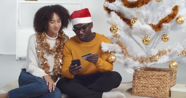 온라인 크리스마스 쇼핑. 인터넷 가게에서 선물을 사는 휴대 전화와 신용 카드를 가진 행복 한 젊은 남자와 곱슬머리 민족 여성, 새해 휴가 할인, 생태계 개념 — 비디오