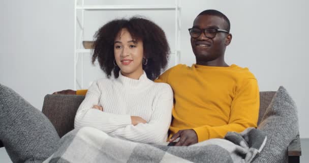Feliz casado casal afro-americano homem e mulher assistindo filmes programa de TV sentado no sofá sofá, descansando em casa. Jovem namorada da família étnica e namorado passar o tempo juntos na sala de estar — Vídeo de Stock