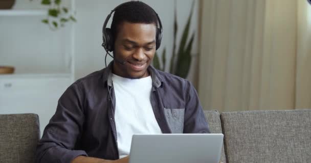 Afro-Amerikaanse man draagt hoofd microfoon en hoofdtelefoon spreekt op webcam antwoorden videogesprek communiceert in online chat verre werk, zwarte man student studies op afstand van huis zitten op de bank — Stockvideo