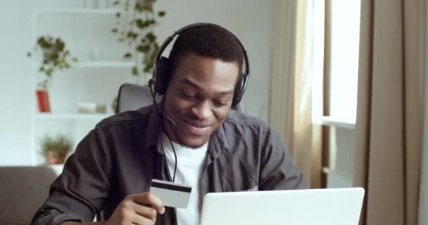 Afro american man in headphones czarny facet student manager składa zamówienie online w sklepie internetowym książki bilety wpisując numery kodu z plastikowej karty kredytowej siedzi przy stole w biurze domu — Wideo stockowe