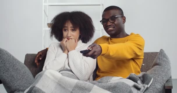 Jovem cara negro e menina americana africana sentado no sofá em casa assistindo filme de terror, mulher de cabelos encaracolados esposa sentindo medo do programa de tv, homem muda de canal com controle remoto — Vídeo de Stock