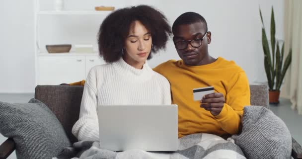 アフリカ系アメリカ人のカップルの夫とメガネと巻き毛の妻は一緒にノートパソコンの予約注文チケットを使用してオンライン購入を行います割引をお楽しみくださいプラスチック銀行カード、電子商取引と販売コンセプト — ストック動画