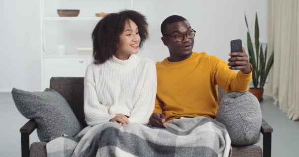 Een paar millennials afro-Amerikaanse familie vrouw en man thuis zitten op de bank doen video chat conferentie met mobiele telefoon praten zwaaien handen hallo aan smartphone camera maakt video online blog — Stockvideo