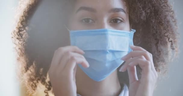 Портрет кучерявої молодої змішаної медсестри, яка щасливо дивиться на камеру, позуючи з посмішкою на обличчі, відчуваючи радість і полегшення після закінчення епідемії вірусу, знімаючи її медичну захисну маску — стокове відео