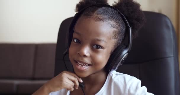 Πορτρέτο της μικρής συναισθηματικής Αφρικής κορίτσι 5 -7 ετών κάθεται στο τραπέζι στο σπίτι μπροστά από φορητό υπολογιστή φοράει μικρόφωνο κεφαλής μιλάει επικοινωνεί online μελέτες εξ αποστάσεως δείχνει την κάμερα με το δάχτυλο — Αρχείο Βίντεο
