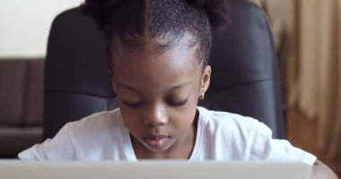 Yoğunlaşmış anaokulu öğrencisi kız çocuğunun portresi dizüstü bilgisayara uzaktan bakarak evden ders çalışıyor, siyah bir çocuğun yakından video dersi izlediği suratında ciddi ifade var.