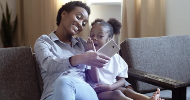 Όμορφη νεαρή Αφρο-Αμερικανίδα μητέρα κάθεται στον καναπέ με αστεία χαριτωμένο κοριτσάκι βγάζοντας φωτογραφίες selfie με κάμερα smartphone. Εθνοτική γυναίκα με μικρό παιδί κάνει φωτογραφίες selfies στο σπίτι δωμάτιο — Αρχείο Βίντεο