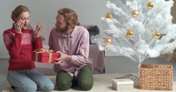 Καυκάσια οικογένεια γιορτάζει το νέο έτος μαζί στο σπίτι, νεαρή ξανθιά γυναίκα κάθεται καλύπτει τα μάτια της με τα χέρια της καυκάσιος γενειοφόρος άνδρας δίνει στη σύζυγό του φίλη χριστουγεννιάτικο δώρο κόκκινο κουτί δώρου — Αρχείο Βίντεο