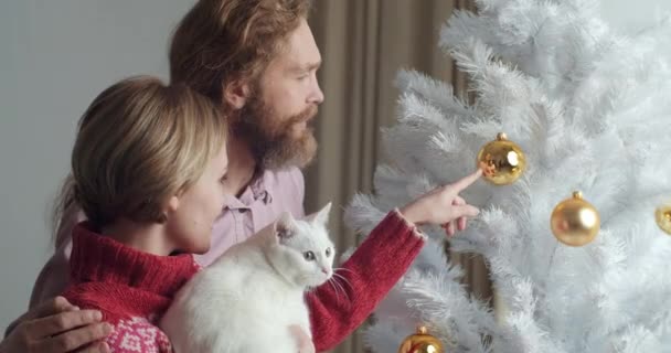 Close-up retrato de casal caucasiano com gato branco seu amado animal de estimação fica abraçando perto da árvore de Natal mostra animal bola amarela decoração de Ano Novo, marido e esposa celebrar férias em aconchegante — Vídeo de Stock