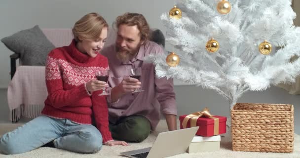Кавказская молодая семья тысячелетий бородатый мужчина и блондинка, сидящие вместе возле елки, пить вино, звон очков, поздравления удаленно с праздником, выступая в Интернете на веб-камеру — стоковое видео