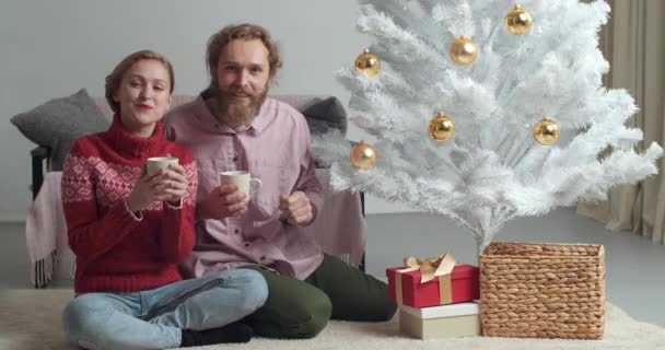 Casal caucasiano esposa e marido sentado em casa perto da árvore de Natal beber de copos de cacau cappuccino café chá vinho quente acenando as mãos para câmera falando desejo felicidade no ano novo 2021 — Vídeo de Stock