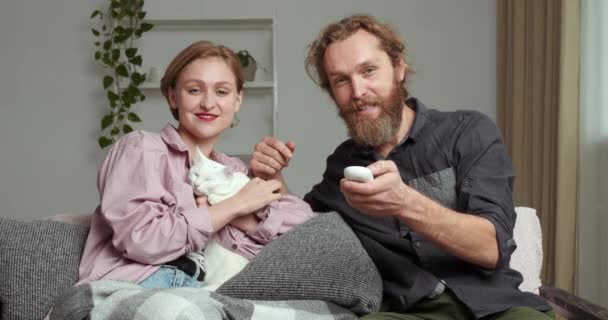 Καυκάσια ζευγάρι ξανθιά γυναίκα και γενειοφόρος άνδρας κάθεται με λευκή χνουδωτή γάτα στον καναπέ στο σαλόνι βλέποντας ταινίες τηλεόραση χαλαρώνοντας στον καναπέ ο σύζυγος αλλάζει κανάλι ενώ η γυναίκα του χαϊδεύει το αγαπημένο του κατοικίδιο — Αρχείο Βίντεο