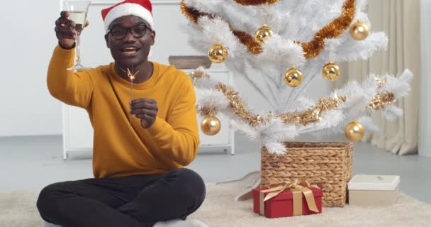 Afro amerykański facet czarny człowiek w okularach i żółty sweter siedzi w pobliżu choinki na podłodze świętując nowy rok 2021 uśmiechnięty podnosi kieliszek szampana trzyma blask w ręce życzy szczęścia — Wideo stockowe