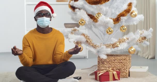 Afro amerykański facet czarny mężczyzna nosi świąteczny kapelusz i medyczna maska ochronna czuje równowagę siedząc w pozycji lotosu na podłodze w domu praktykując jogę medytując z zamkniętymi oczami w Nowy Rok wakacje — Wideo stockowe
