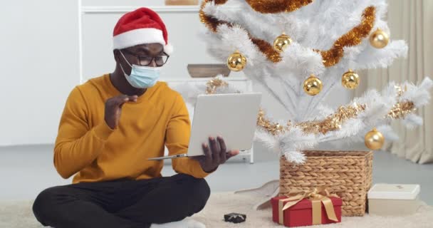 Afro amerykański facet nosi kapelusz jak Święty Mikołaj i medyczna maska ochronna siedzi w pobliżu choinki używa laptopa do zdalnej komunikacji gratuluje rodzinie mówi do kamery podczas epidemii — Wideo stockowe