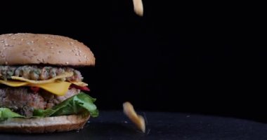 Fast food konsepti. Patates kızartması düşmek siyah masaya, taze çizburgerin yanında uçmak, nefis bir lezzet, karanlık arka planda iştah açıcı bir hamburger, açlık hissi uyandıran bir resim.