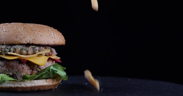 Концепция быстрого питания. Падающий картофель фри жареный картофель падает на черный стол рядом со свежим чизбургером, небесное наслаждение, аппетитный гамбургер на темном фоне близко, картина вызывает голод — стоковое видео