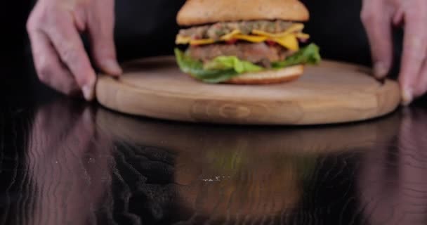 Chef hombre de cerca de las manos masculinas sostiene fresca hamburguesa en bandeja redonda de madera sobre fondo negro de la cocina. Bollo, chuleta frita, lechuga verde jugosa, tocino crujiente, rebanada de queso y tomate, comida poco saludable — Vídeos de Stock