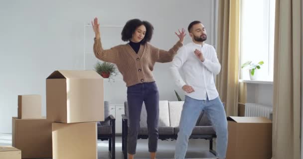 Jeune couple multinational multiracial dansant dans une nouvelle maison célébrant déménagement famille location appartement se sentant heureux déménagement à la musique se réjouissant entouré de boîtes en carton de colis dans le salon — Video