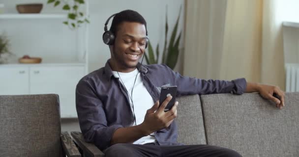 Preto cara afro americano homem sentado no sofá em casa no escritório descansando com fones de ouvido ouvir música assistindo vídeo online na internet através de celular smartphone gadget apreciando música dançando cabeça — Vídeo de Stock