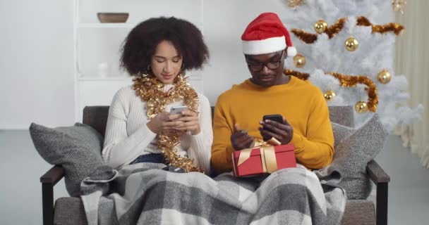 Afroamerykanie para przyjaciele siedzi na kanapie w świąteczne stroje patrząc na telefony komórkowe przeglądanie na czacie w internecie facet pokazuje dziewczyna muzyka wideo dziewczyna taniec porusza głową do muzyki — Wideo stockowe