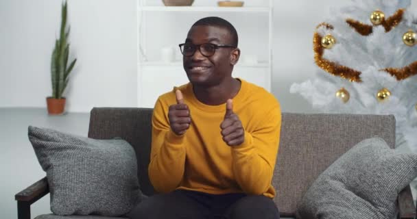 Portrait eines Afroamerikaners mit Brille und gelbem Pullover sitzt auf dem heimischen Sofa in der Nähe von Weihnachtsbaum Talkshows wie Daumen hoch Geste der Zustimmung für positive Einstellung, Blick aus der Web-Kamera — Stockvideo