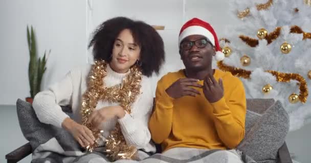 Festa di Capodanno a casa ragazza afro-americana e ragazzo nero in costumi e decorazioni natalizie seduti insieme sul divano a ballare, fidanzato che canta canzoni rap alla musica, fidanzata che si muove ritmicamente — Video Stock