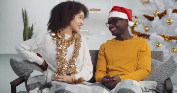 Η σγουρομάλλα φιλενάδα σύζυγος αδελφή κορίτσι δίνει στον άντρα της φίλο μαύρο άντρα Αφροαμερικανό αγόρι Χριστουγεννιάτικο δώρο κουτί σε κόκκινο περιτύλιγμα συγχαίρει για τις διακοπές του νέου έτους στο σπίτι κάθεται στον καναπέ — Αρχείο Βίντεο