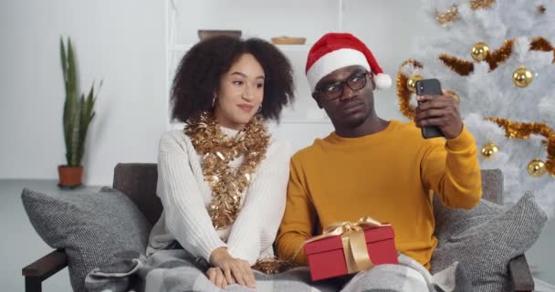 幸せな若いアフリカ系アメリカ人のカップルはクリスマスツリーの近くのソファに座ってビデオメッセージを記録します。クリスマスに携帯電話のカメラの前で手を振っている暖かい天気の2人の陽気な人々 — ストック動画