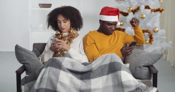 Afro αμερικανικό ζευγάρι κάθεται σε καναπέ τα Χριστούγεννα κοιτάζοντας προς διαφορετικές κατευθύνσεις χρησιμοποιούν τηλέφωνα κινητό έξυπνο τηλέφωνο παίζουν κουβέντα σε απευθείας σύνδεση έχουν μυστικά αισθάνονται βαριούνται ζουν σε φιλονικία, εθισμού έννοια πρόβλημα — Αρχείο Βίντεο