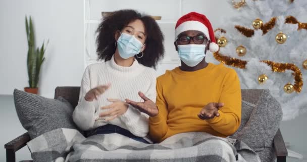 Webcam vista de la familia de raza mixta en máscaras de protección médica sentado en la sala de estar cerca del árbol de Navidad saludando hablando en el chat de vídeo durante el aislamiento de cuarentena, año nuevo 2021 en coronavirus — Vídeos de Stock