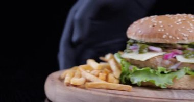 Yandan kesilmiş, lastik lateks eldivenli erkek elleri masaya ahşap tahta, fast food, yağlı hamburger, peynirli, sebze soslu salata ve patates kızartması koyuyor.