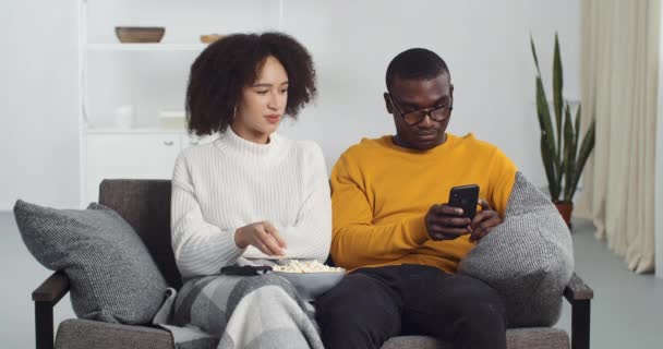 Mladý kudrnatý manželka afro americká dívka a smíšené rasy černošky manžel chlap s brýlemi sedí spolu na dece na pohovce v útulném obývacím pokoji, jíst popcorn mluvit při pohledu na obrazovce mobilního smartphonu — Stock video